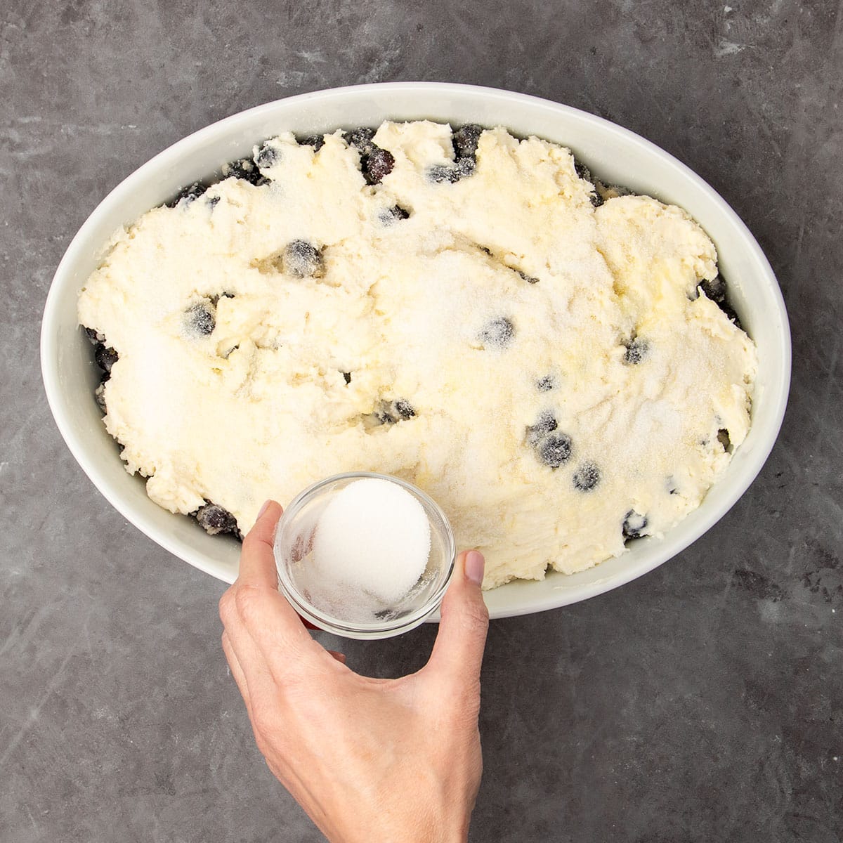 hand holding very small glass dish sprinkling white granulated sugar on top of topping in white dish