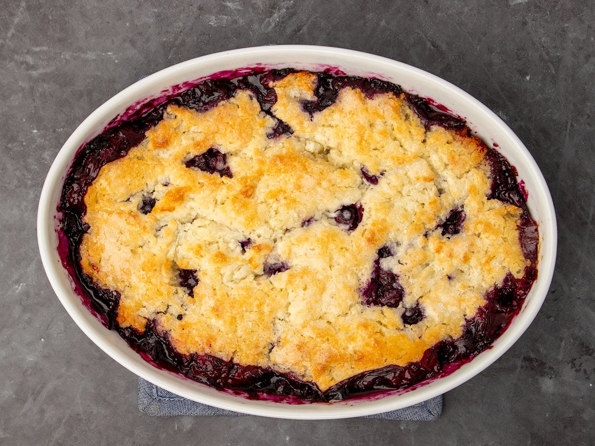 overhead image of baked bubbling blueberry cobbler filling with baked golden brown pastry topping in white casserole dish
