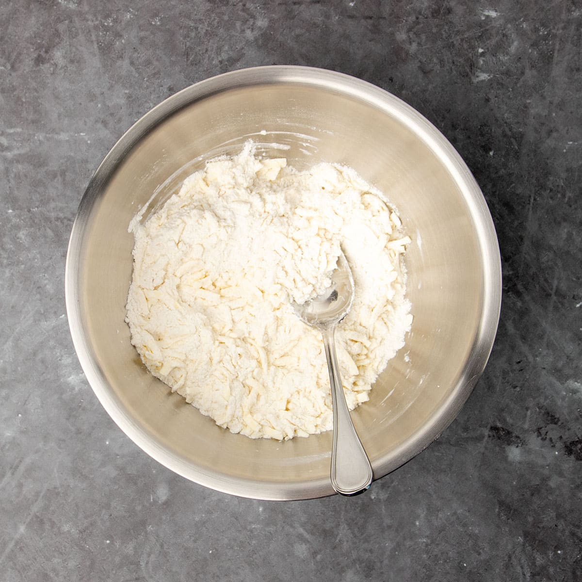 same mixing bowl with grated light yellow butter shreds mixed in to flour with large metal mixing spoon