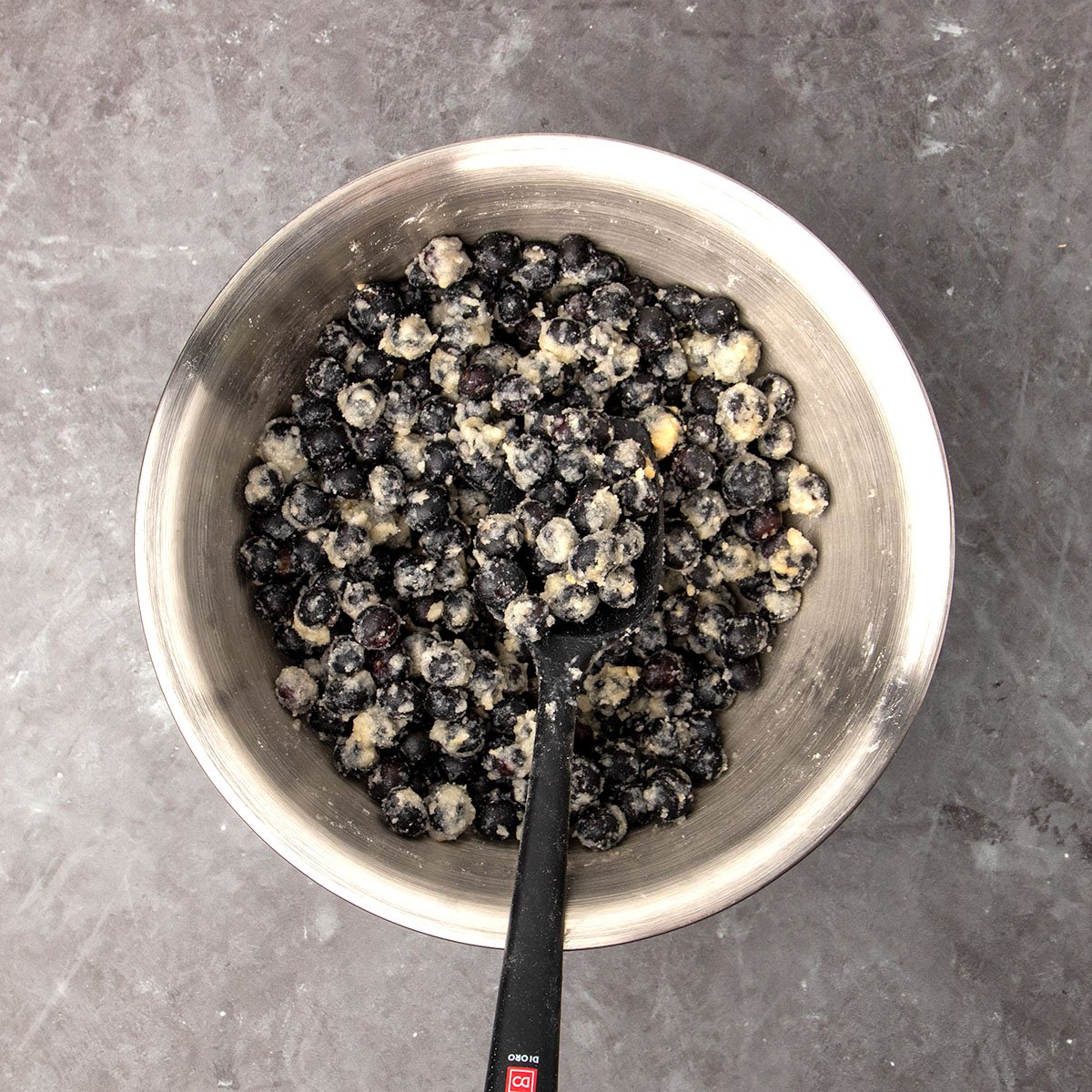 Large round metal mixing bowl with blueberries mixed with melted butter and flour with black silicone spatula