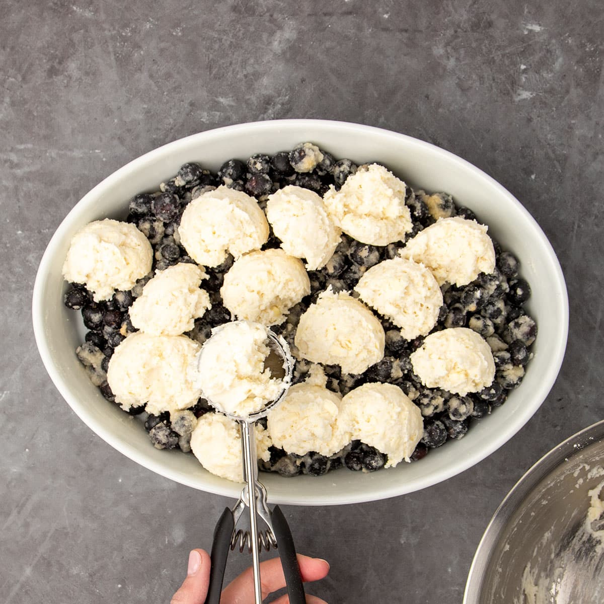 scoops of white topping mixture on top of blueberries in white dish with hand holding metal ice cream scoop with some more topping mixture in bowl of scoop
