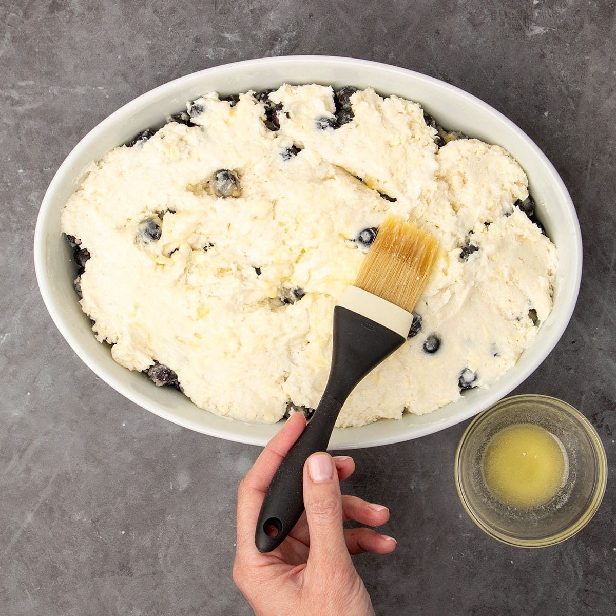 hand holding pastry brush with black handle brushing melted yellow butter on top of topping in white dish