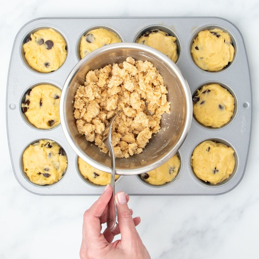 same crumble mixture in bowl now chilled and lighter brown in color with hand holding a fork breaking it up into chunks, over metal muffin tin with batter in wells