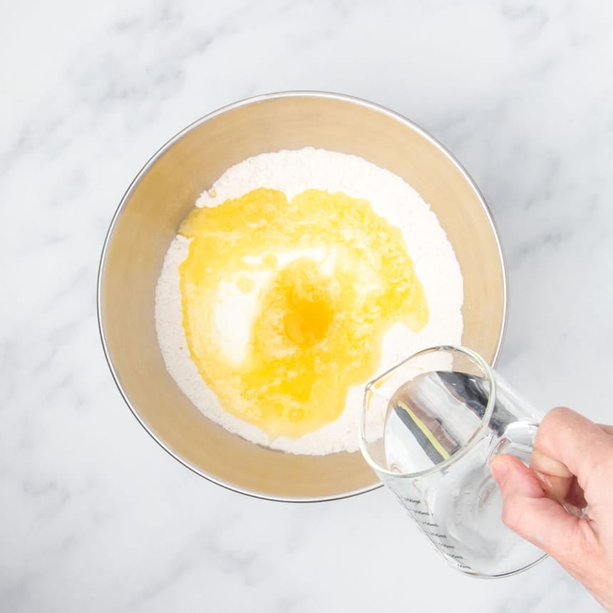 hand holding small pitcher pouring water into same mixing bowl with yellow egg and melted butter and white yogurt on top of flour in bowl