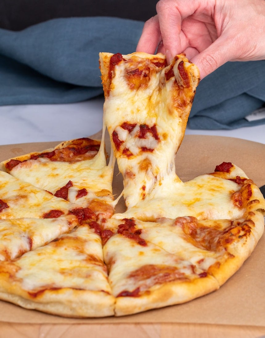 fingers of hand lifting triangular shaped slice of pizza from a pie cut into 8 slices on brown paper with blue cloth