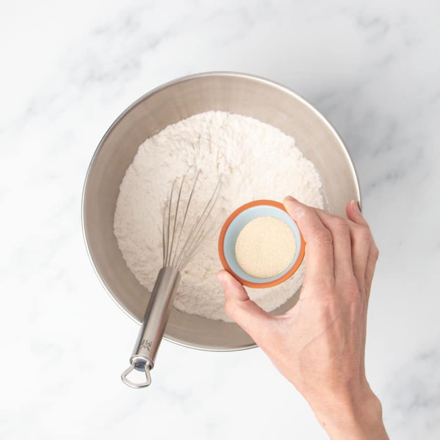 hand holding small blue bowl with light brown yeast granules over a large metal round mixing bowl with flour and a wire whisk