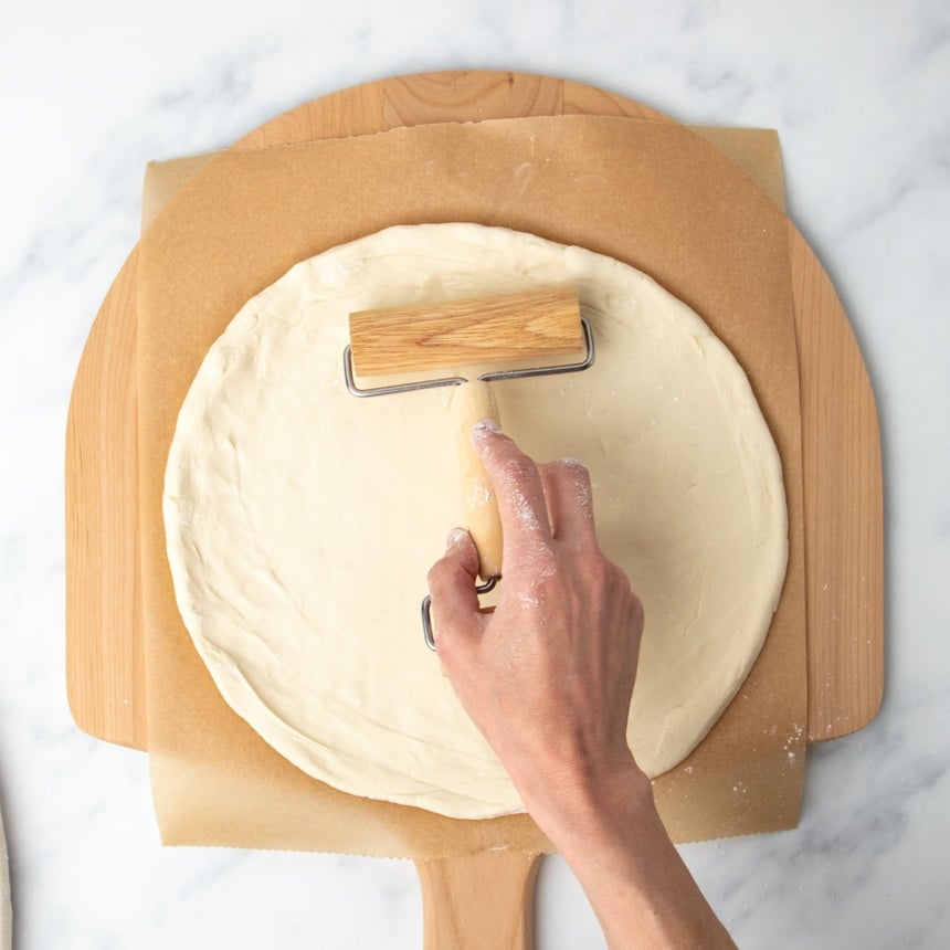hand using small light blond wood pizza roller to press same dough into a large, smooth round, with dough now on brown paper on a blond wood pizza peel