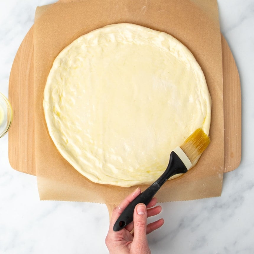 hand using pastry brush with black handle to brush olive oil on edge of raw round of pizza dough
