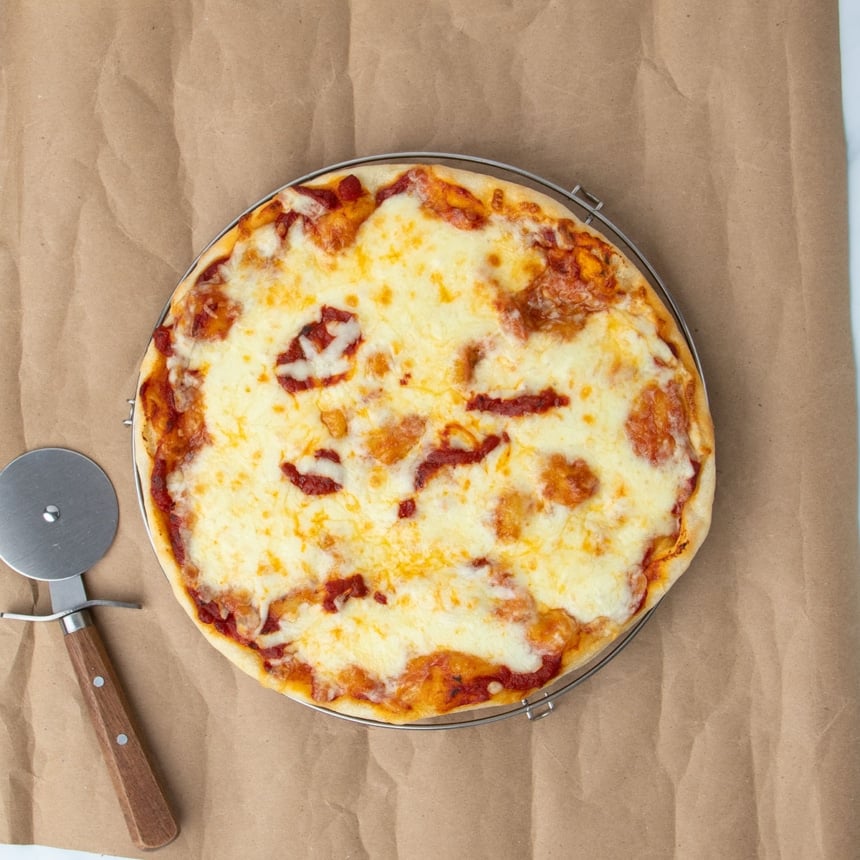 fully baked gluten free pizza round with red sauce peeking through melted off white cheese on brown paper with brown handled metal pizza wheel, all on brown kraft paper