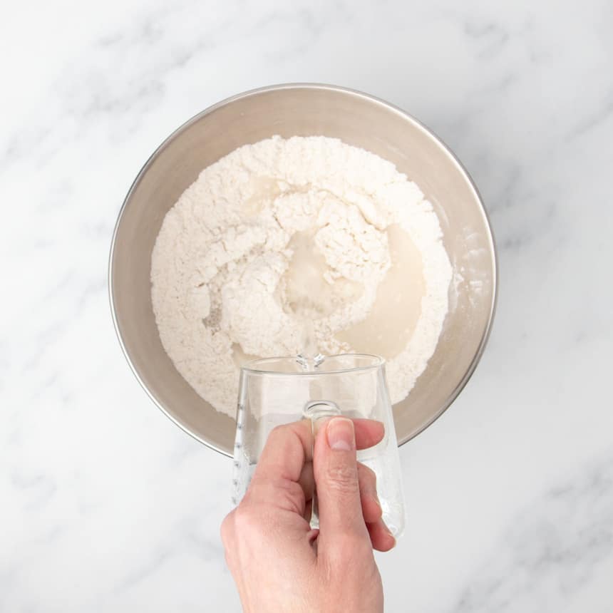 hand pouring water from a small glass pitcher into the same large metal mixing bowl with whisked flour and other dry ingredients