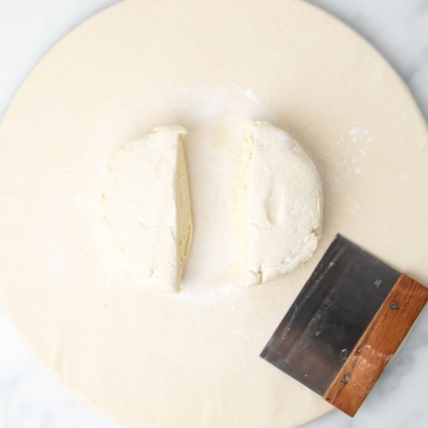 same pizza dough on light tan canvas covered board sliced in half with metal bench scraper with brown wood handle