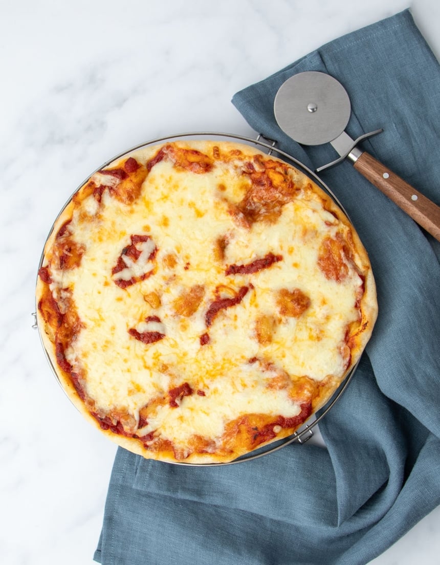 round wire rack below fully baked gluten free pizza with red tomato sauce, melted off white cheese, with blue cloth and metal pizza wheel with brown handle
