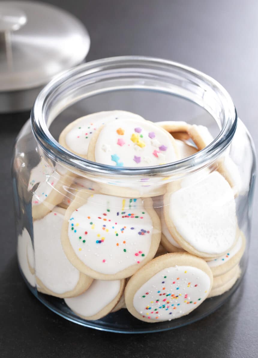 A large glass jar full of round pale cookies with white frosted and colored nonpareils