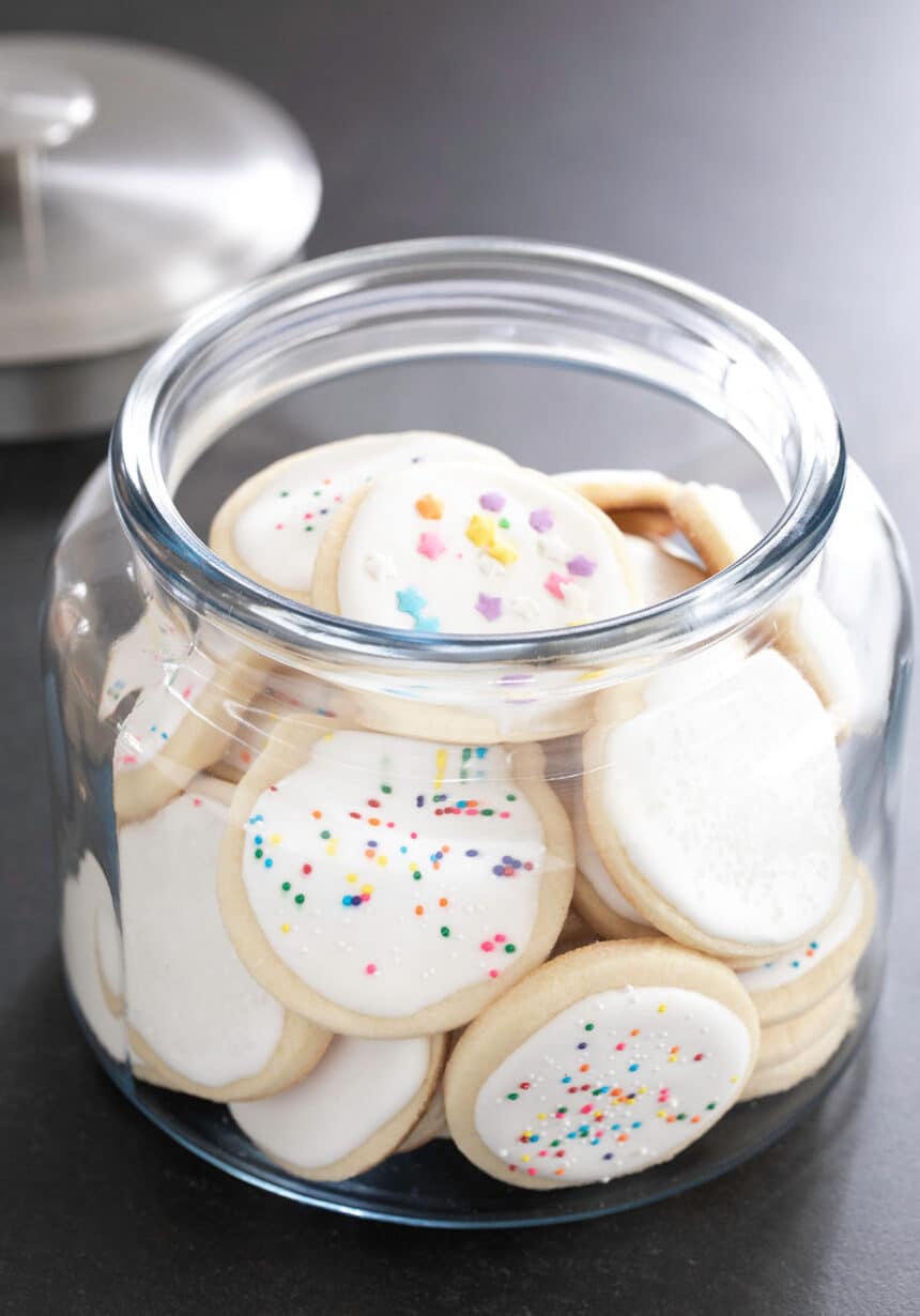 A large glass jar full of round pale cookies with white frosted and colored nonpareils