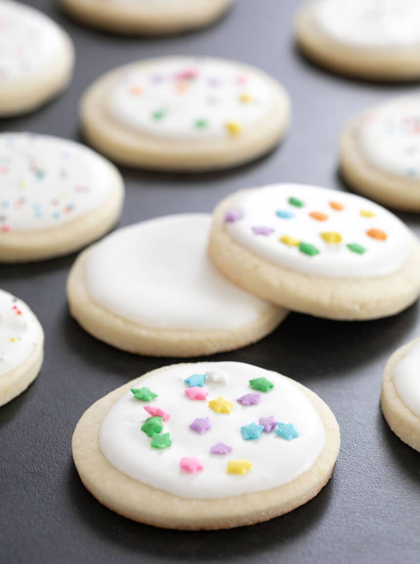 Close up of round pale cookies with royal icing and star sprinkles on a dark surface.