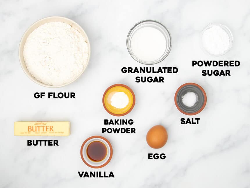 ingredients for gluten free sugar cookies in small bowls on marble surface with black block letters with name of each ingredient