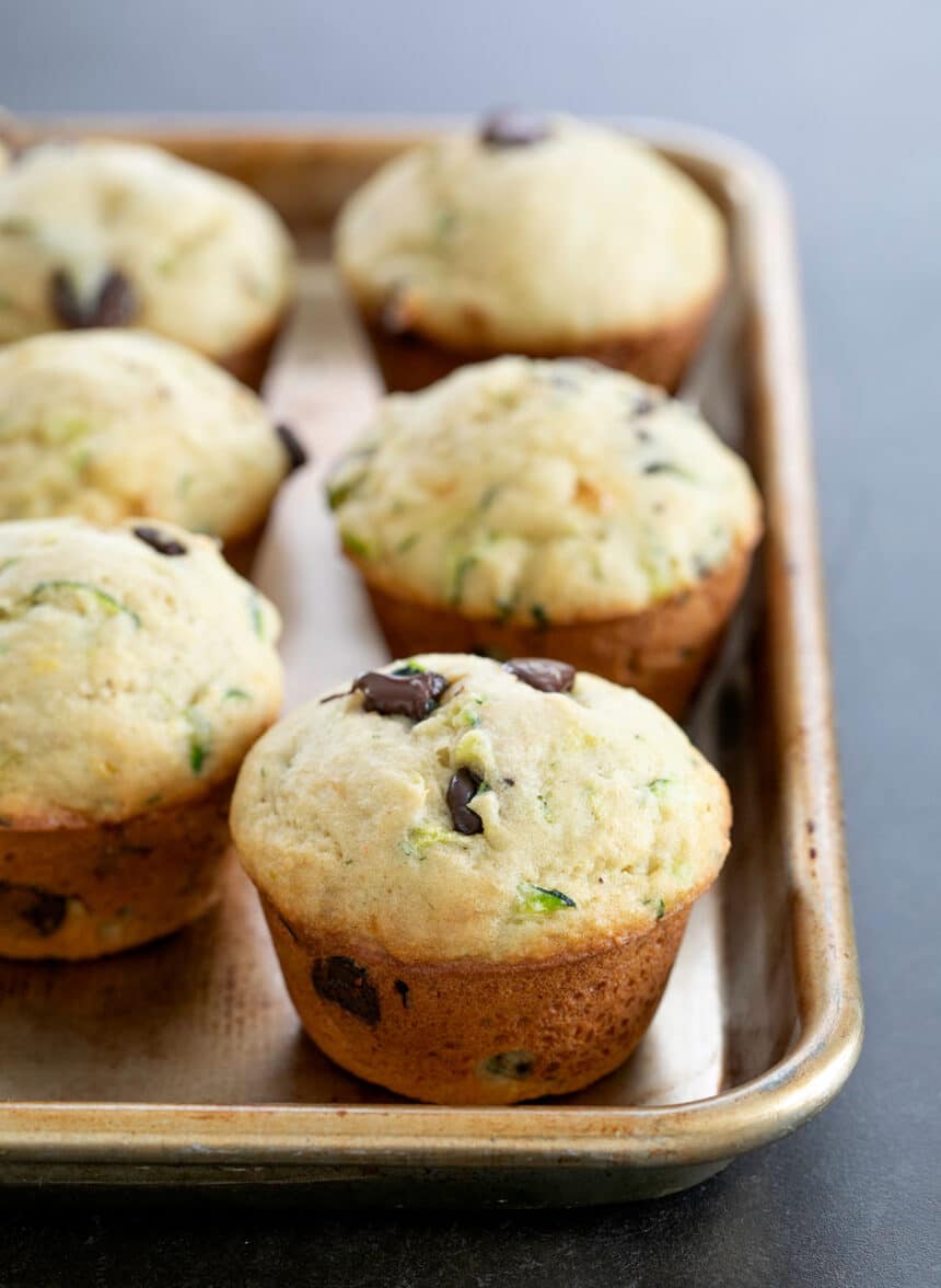 6 light brown muffins with green zucchini flecks and dark brown chocolate chips on a small rimmed metal baking sheet