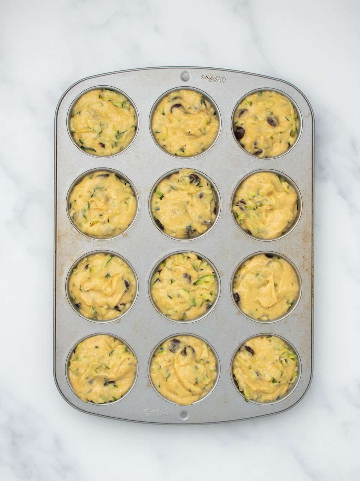 Standard metal 12-cup muffin tin with each well filled with light yellow raw muffin batter.