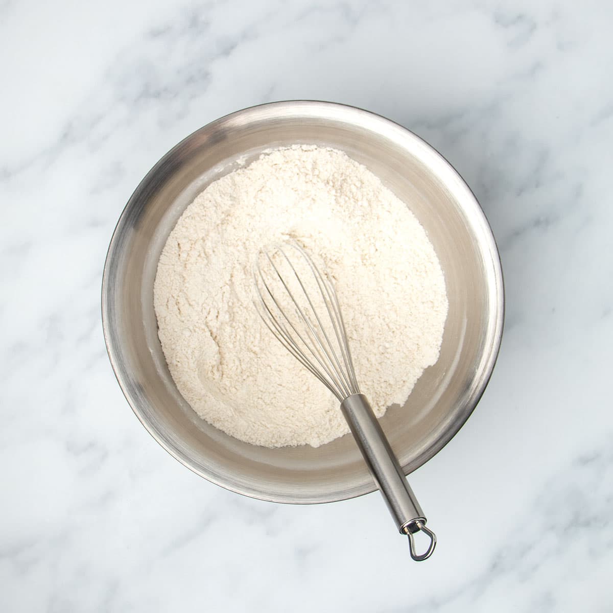 Round metal mixing bowl with white flour mixture and metal wire whisk.