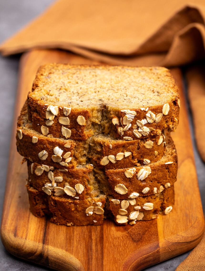 stack of 4 slices of banana bread with old fashioned oats baked onto edge on brown wood paddle board with brown cloth in background