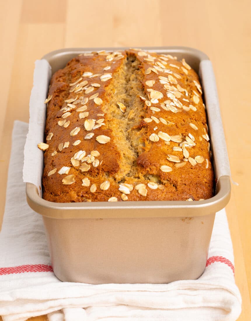 dark brown bread with old fashioned oats baked on top in golden loaf pan lined with white paper on white cloth with red stripe