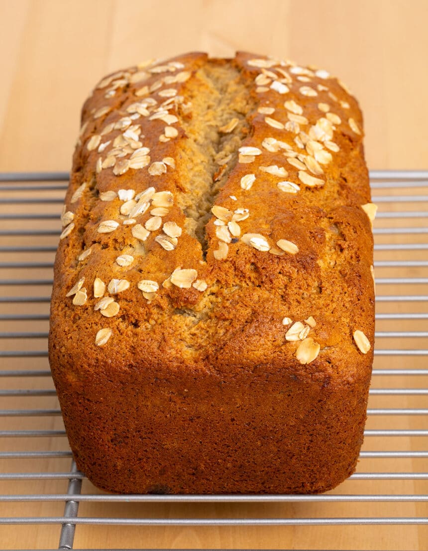 dark brown loaf with oats on top and split down center on light metal cooling rack on top of blond wood surface