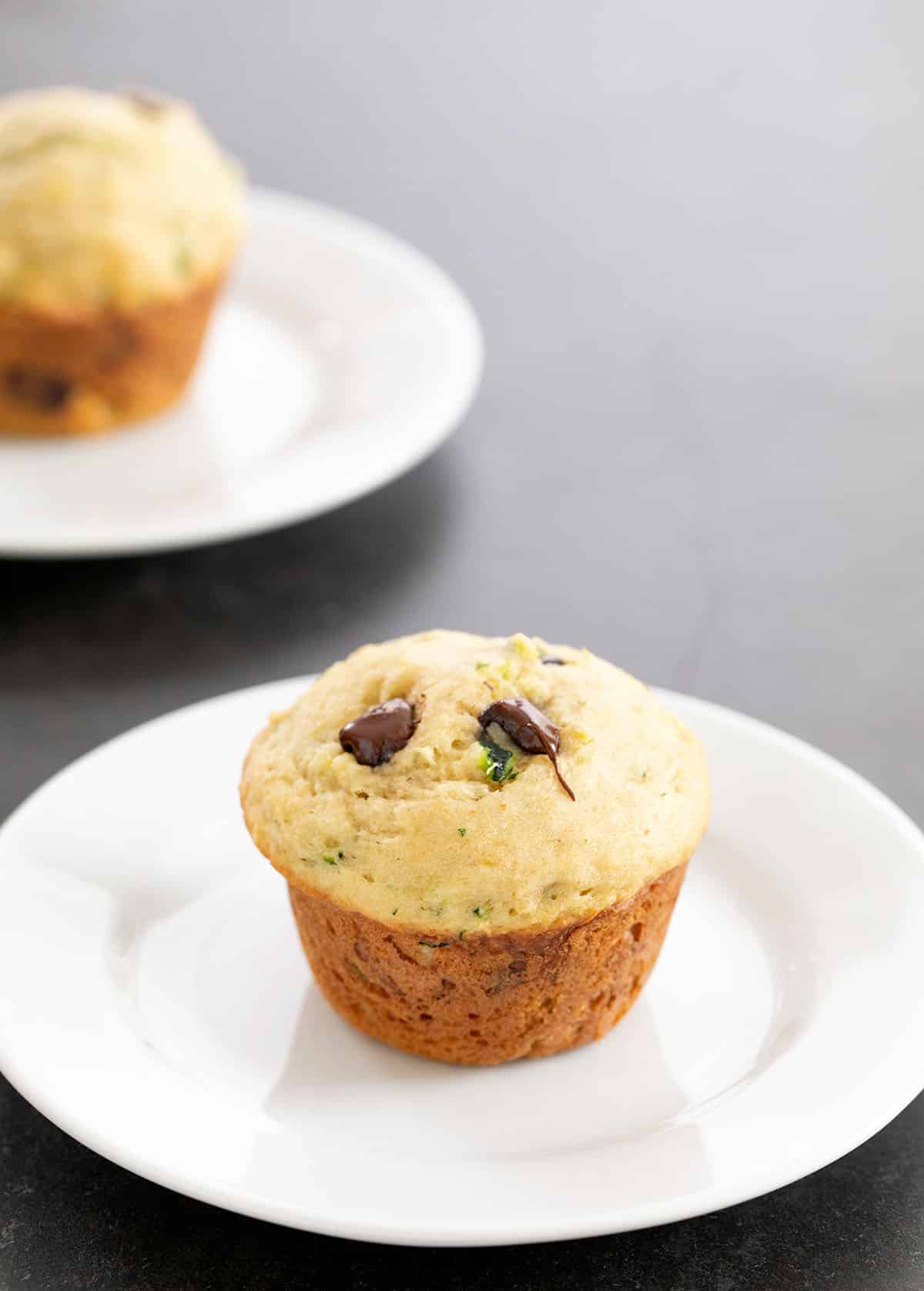 Two zucchini muffins each on a small white plate on a gray surface.