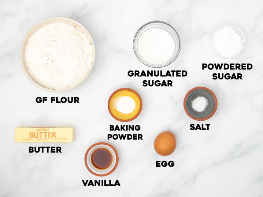 ingredients for gluten free sugar cookies in small bowls on marble surface with black block letters with name of each ingredient