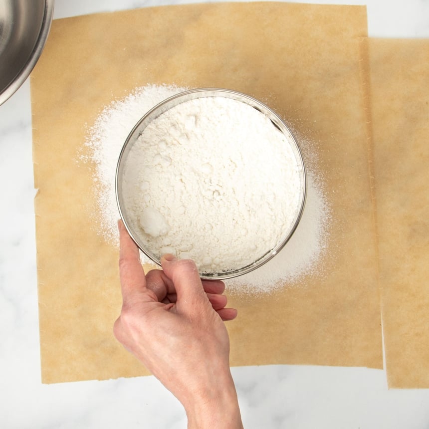 hand holding round metal mesh sieve with white flour over brown sheet of parchment paper