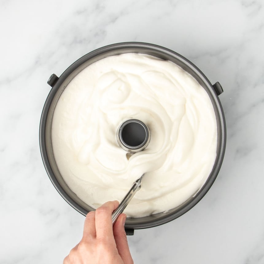 white soft angel food cake batter in black metal tube pan with hand holding knife in mixture