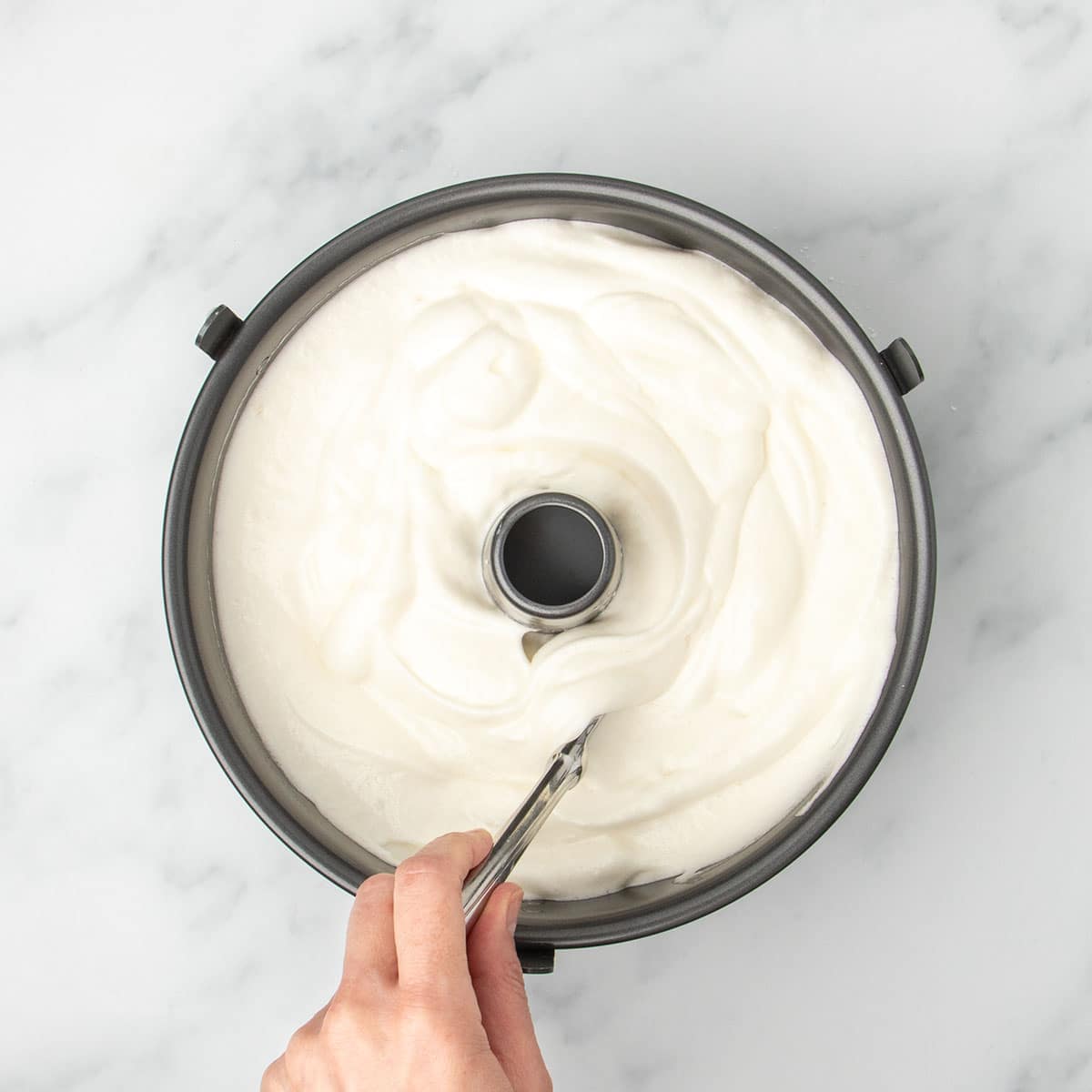white soft angel food cake batter in black metal tube pan with hand holding knife in mixture