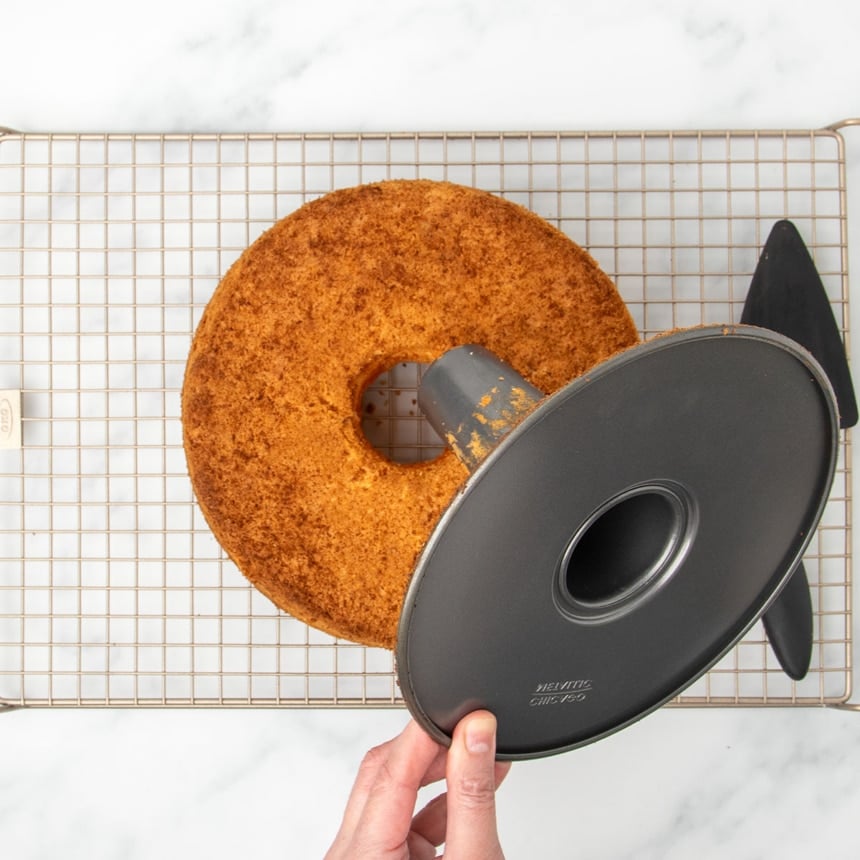 fingers lifting bottom of tube pan out of browned angel food cake on wire rack