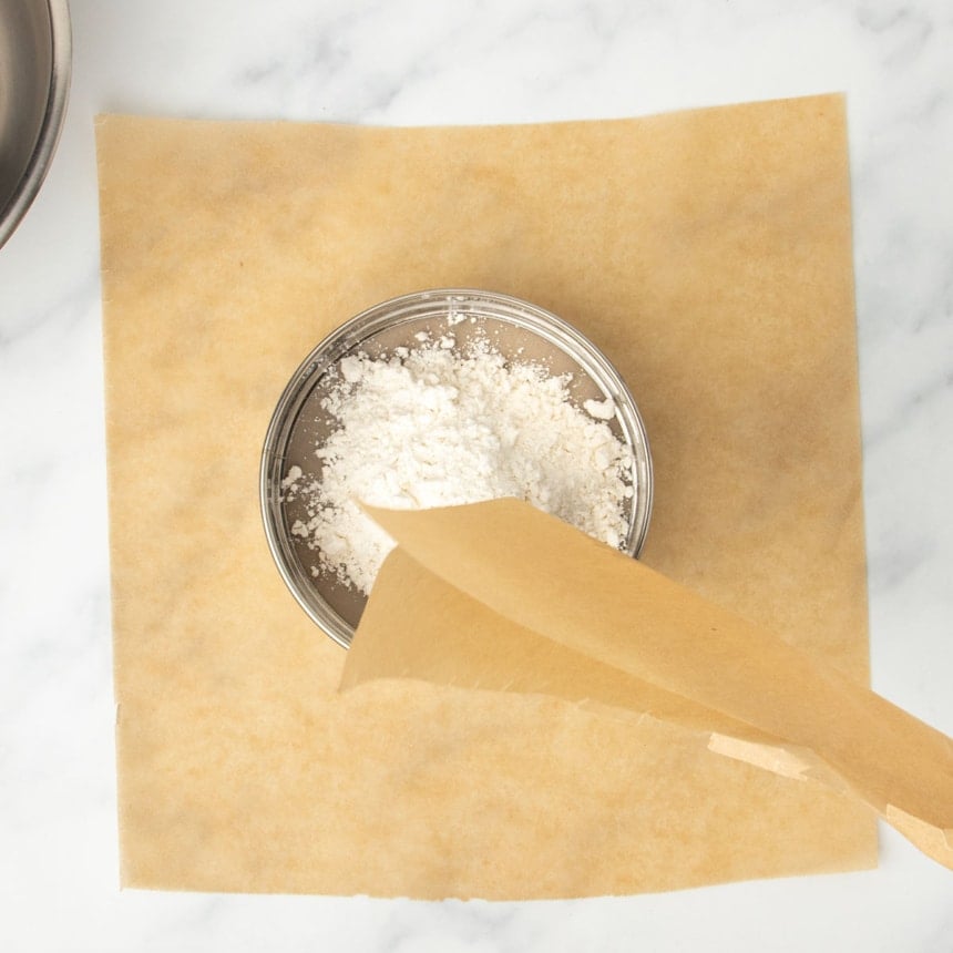 flour being transferred from brown paper folded in half into round metal mesh sieve on top of another sheet of brown paper