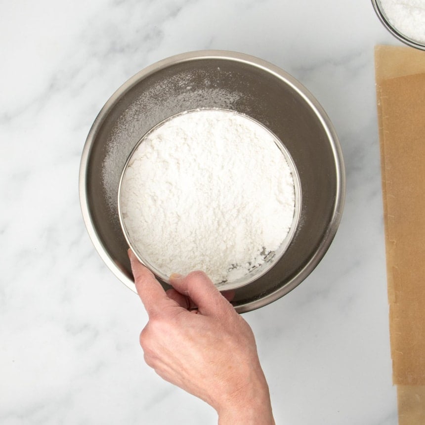 hand holding round metal mesh sieve with white flour over round metal mixing bowl