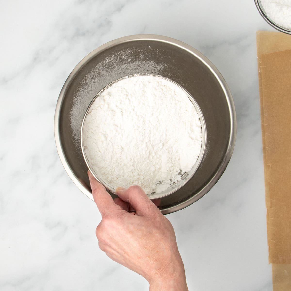 hand holding round metal mesh sieve with white flour over round metal mixing bowl