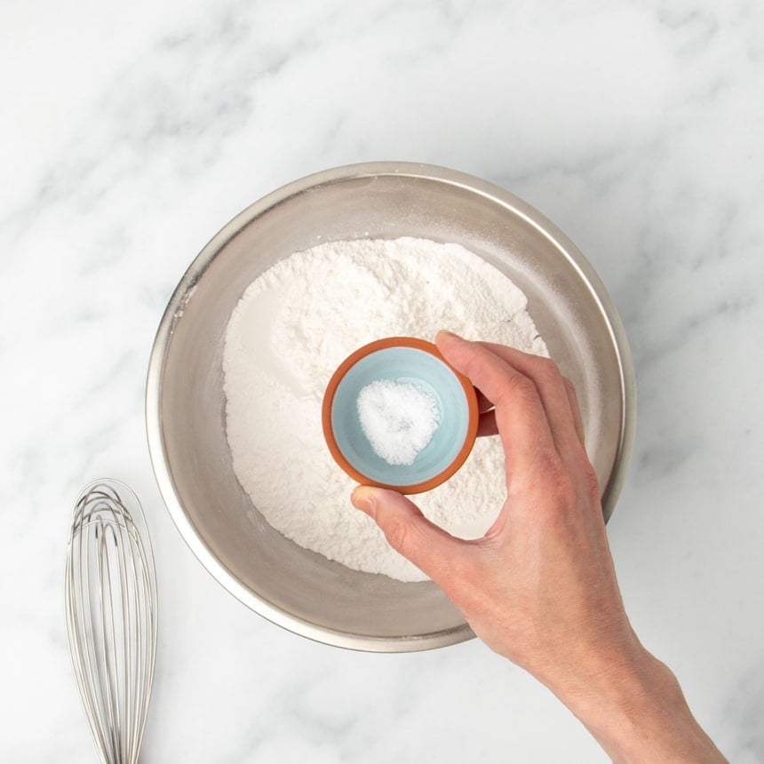 hand holding small blue bowl with salt over metal mixing bowl with flour