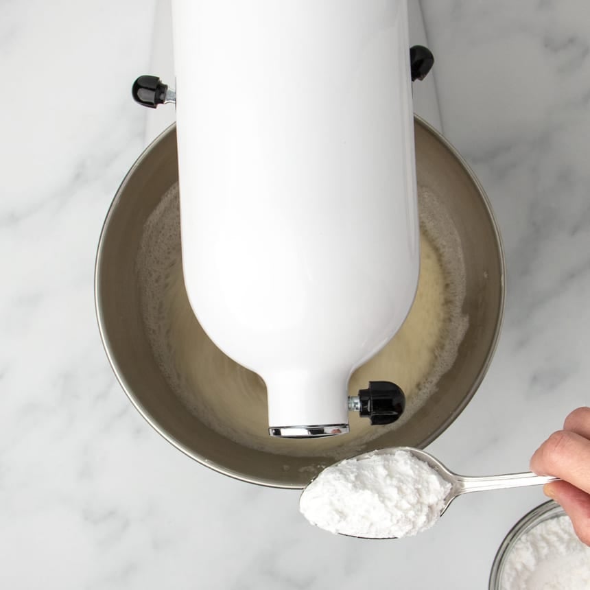 hand holding metal mixing spoon with white flour mixture next to white stand mixer beating egg whites