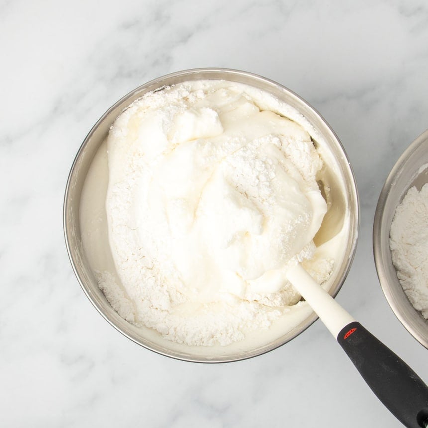 same mixing bowl with white flour mixture being folded into white meringue with white silicone spatula with black handle