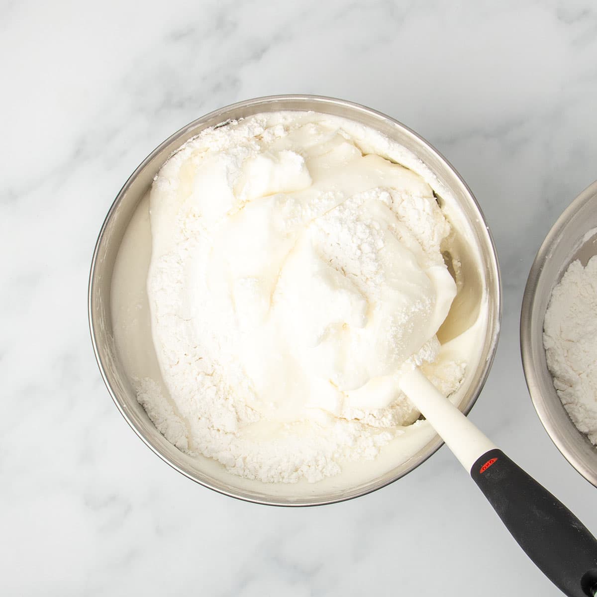 same mixing bowl with white flour mixture being folded into white meringue with white silicone spatula with black handle