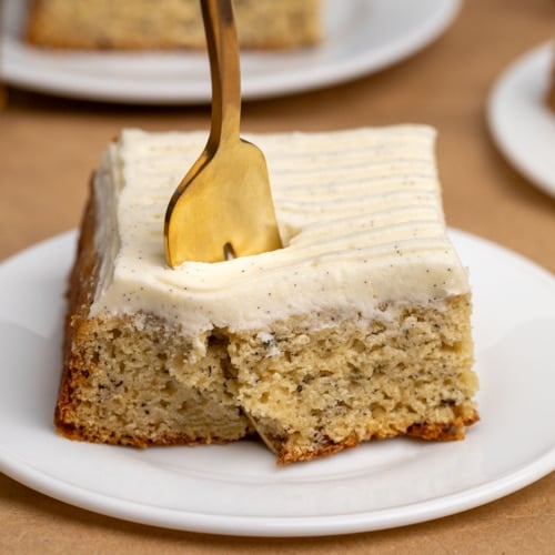 gold fork piercing yellow square of cake with white frosting on small white plate