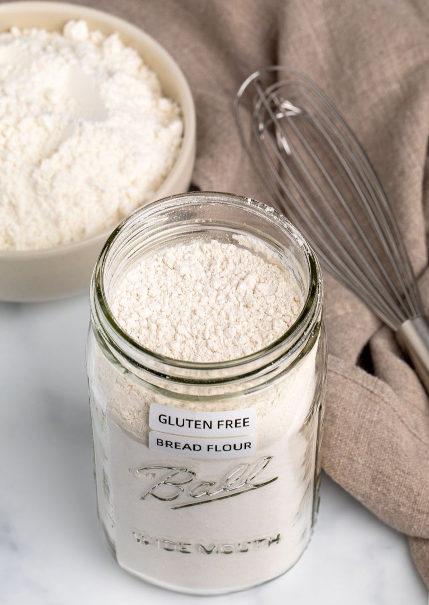 tall wide mouth Ball glass canning jar with white powder and white labels stating "gluten free bread flour" in black writing on marble surface with wire whisk, tan cloth, and small bowl with more flour in background