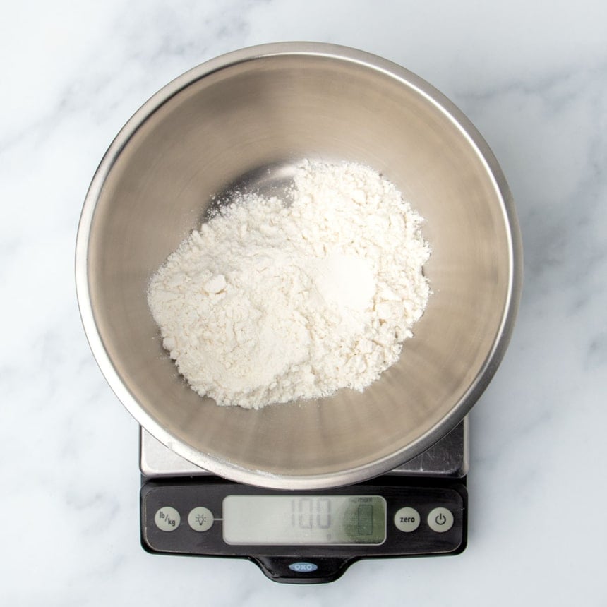 small round metal mixing bowl with white powder on a digital kitchen scale reading 100 grams