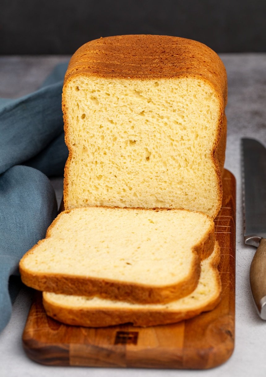 tall partially sliced loaf of bread with a yellow crumb and a brown crust on a narrow dark brown wood cutting board with a blue cloth and metal bread knife with a brown wood handle