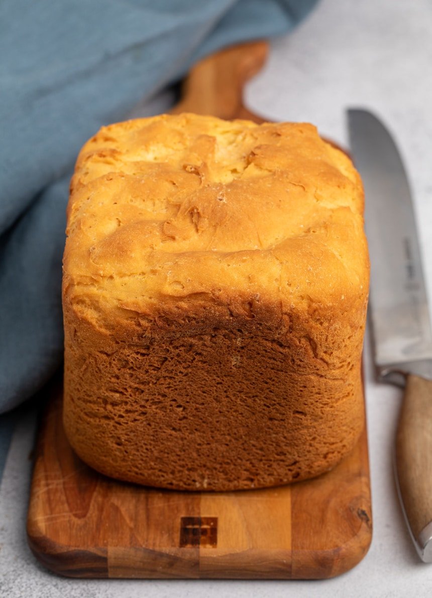whole brown crusted loaf of bread on the same brown cutting board with blue cloth and bread knife