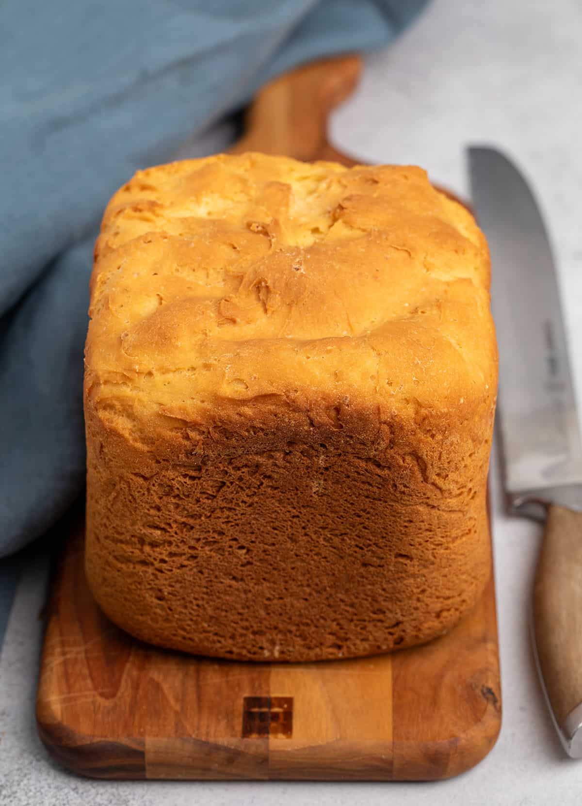 whole brown crusted loaf of bread on the same brown cutting board with blue cloth and bread knife