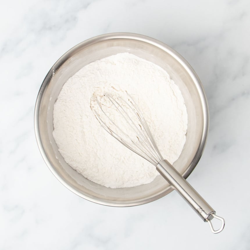 medium round metal mixing bowl with white flour and large metal wire whisk on marble surface