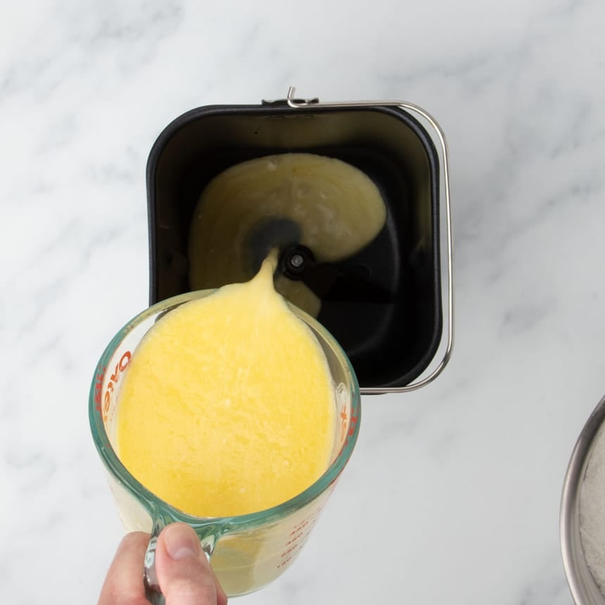 fingers holding 2 cup glass measuring cup pouring yellow mixture into baking pan for bread machine