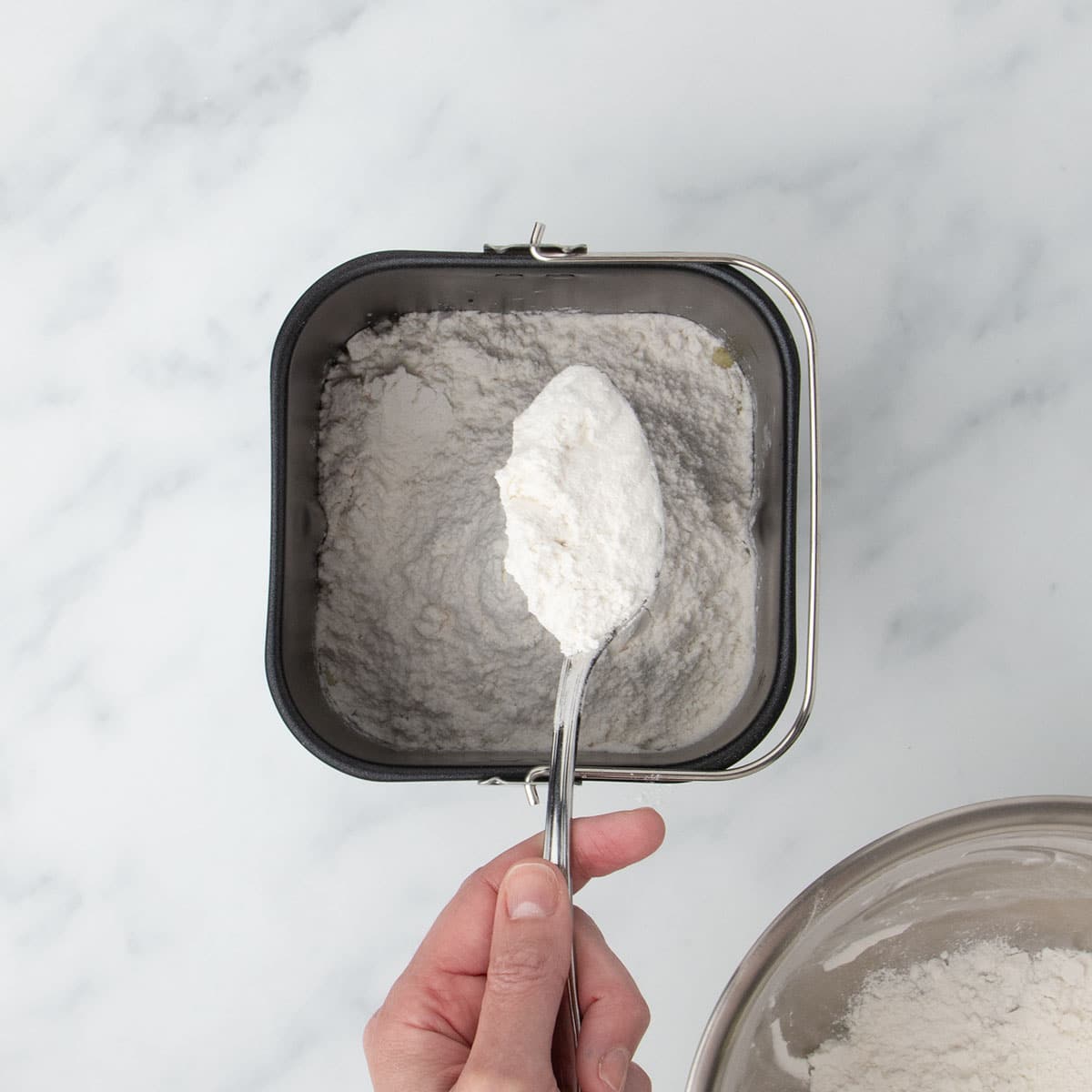 hand holding large metal mixing spoon filled with white flour mixture turning flour over into black bread machine baking pan with more flour