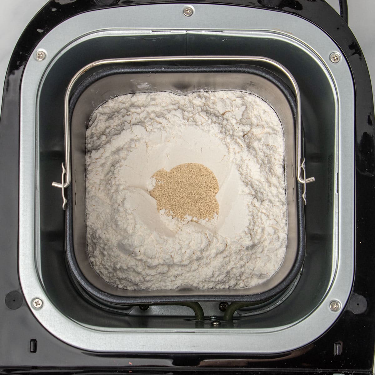 same baking pan with flour on top and instant yeast granules in small hole in center of flour in baking pan in bread machine