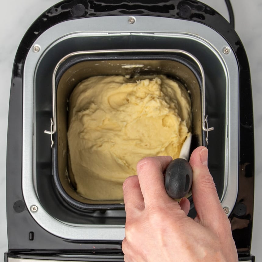 hand holding white silicone spatula with black handle scraping sides of bread machine baking pan with soft raw yellow bread dough